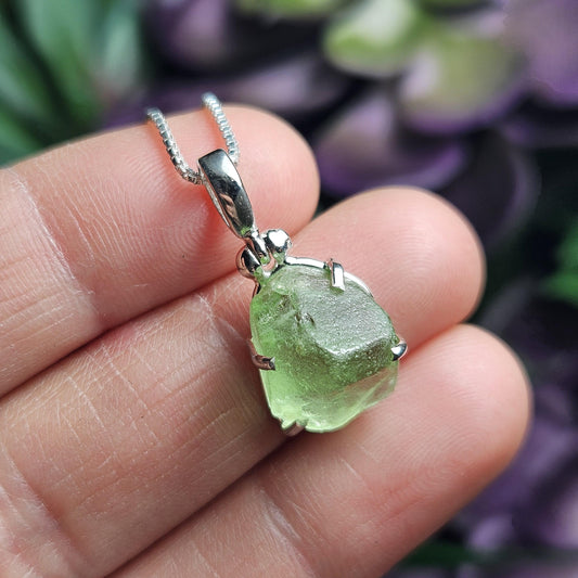 May include: A silver chain necklace with a green gemstone pendant. The pendant is a rough, irregular shape and is set in a silver bezel. The gemstone is a light green color and has a slightly translucent appearance.