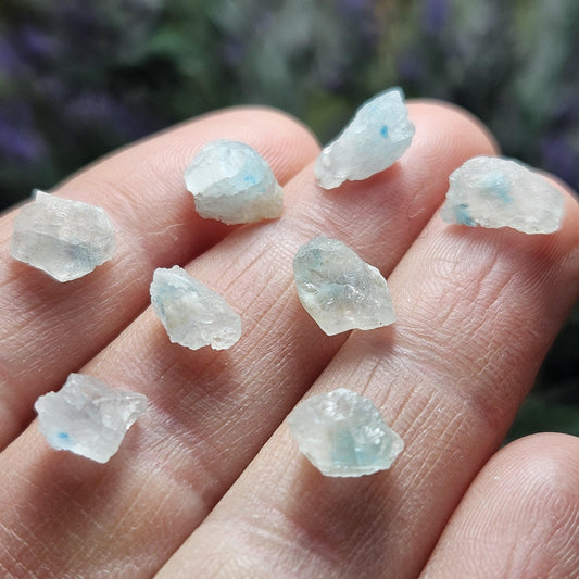 May include: Close-up view of eight small, tumbled stones.  The stones are predominantly translucent white or very light blue, with flecks of darker blue dispersed throughout. They exhibit a rough, natural texture and irregular shapes, characteristic of raw, uncut gemstones. These small, rough, natural stones are ideal for jewelry making, crystal healing, or decorative purposes.