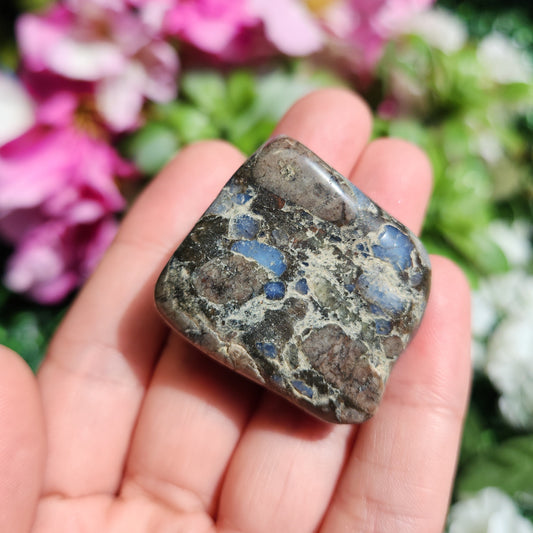 May include: A dark gray, brown, and blue tumbled stone with a smooth, polished surface. The stone has a speckled pattern with blue and white inclusions.