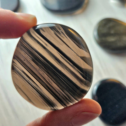 May include: A smooth, round, polished stone with a black and brown striped pattern. The stone is translucent and has a glossy finish.
