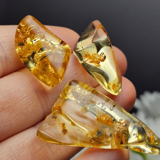 May include: Three pieces of amber with inclusions. The amber is a light yellow color and is transparent. The inclusions are small and dark brown. The amber is shaped like a triangle, a rectangle, and a teardrop.