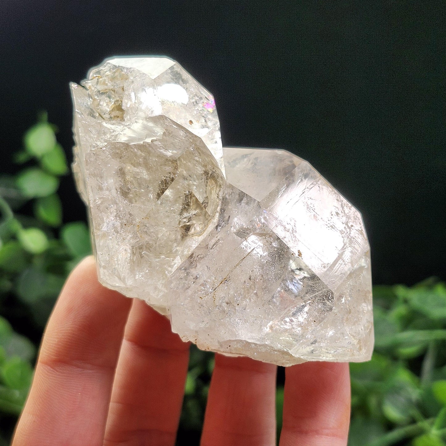 May include: A clear quartz crystal cluster with multiple points. The crystal has a rough, natural texture and a translucent appearance, with subtle internal inclusions. The quartz is held in a hand against a dark green background.