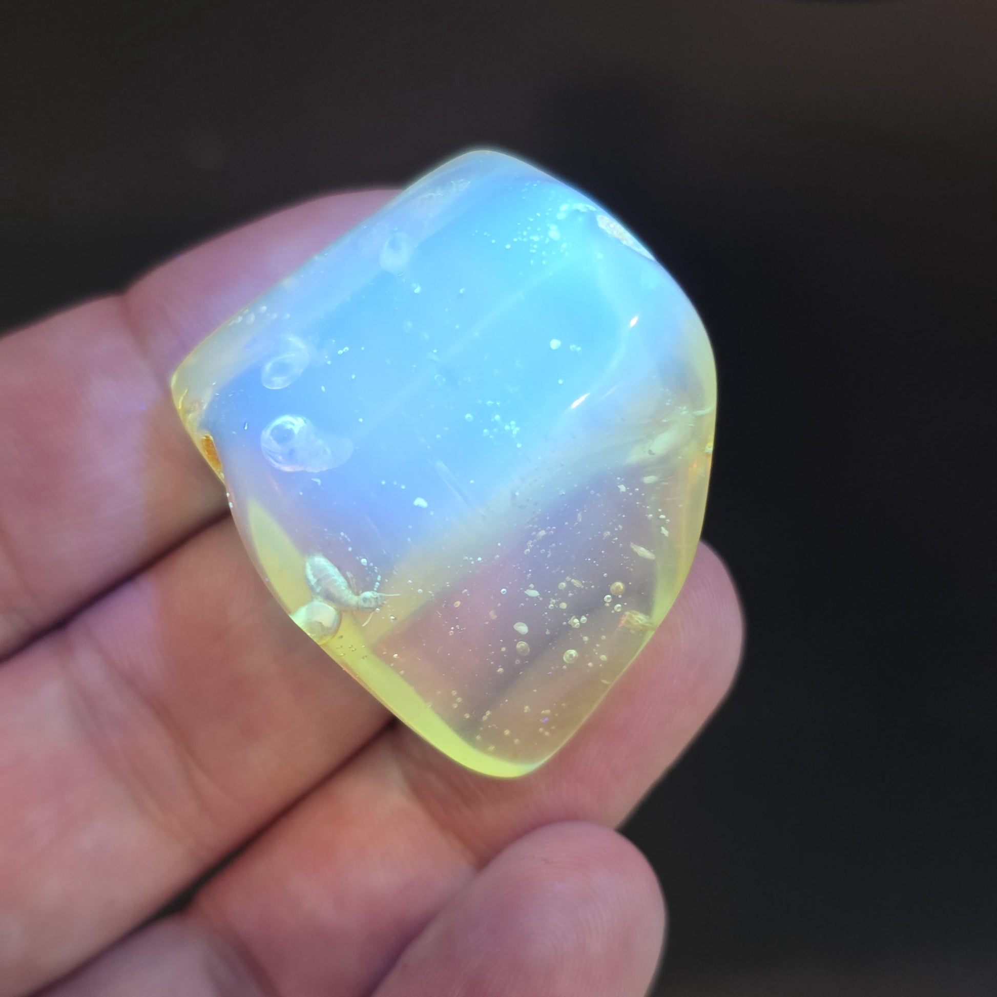 May include: A translucent, polished gemstone with a rounded shape. The stone has a pale yellow hue with internal blue and white iridescent patterns. The gemstone is held in a hand, with a dark background.