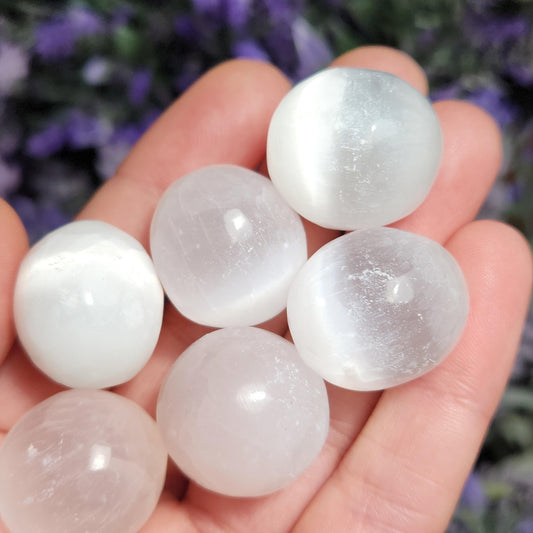 May include: Six smooth, white selenite spheres, also known as moonstone, in various sizes, are held in a hand. The spheres are polished and have a translucent, milky white appearance.
