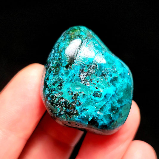 May include: A polished, rounded gemstone with a vibrant turquoise and teal color palette. The stone has dark green and black inclusions, creating a textured appearance. The gemstone is held in a hand against a black background.