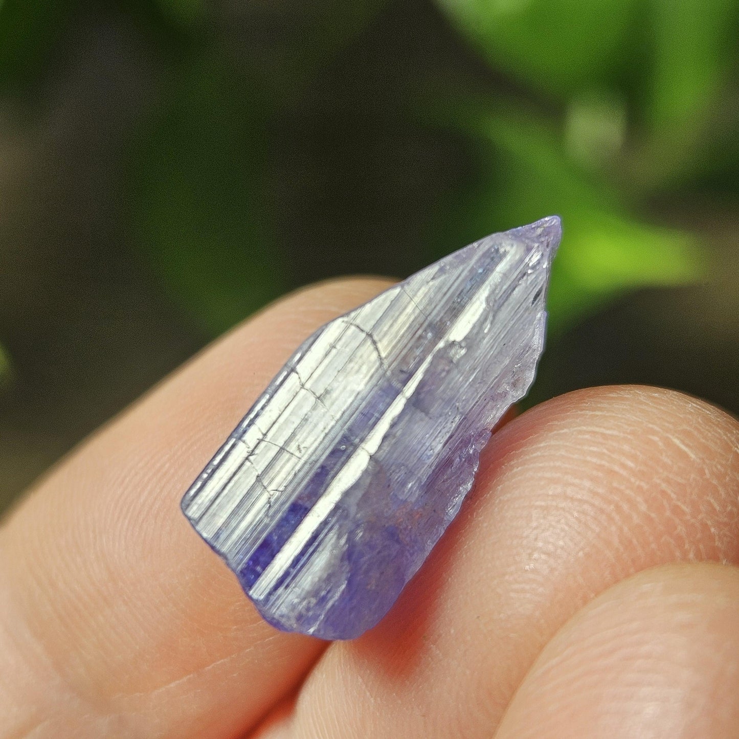 May include: A small, elongated, light purple crystal with a pointed tip. The crystal has a translucent quality with visible internal striations and a subtle sheen. The background is blurred, suggesting a natural setting.