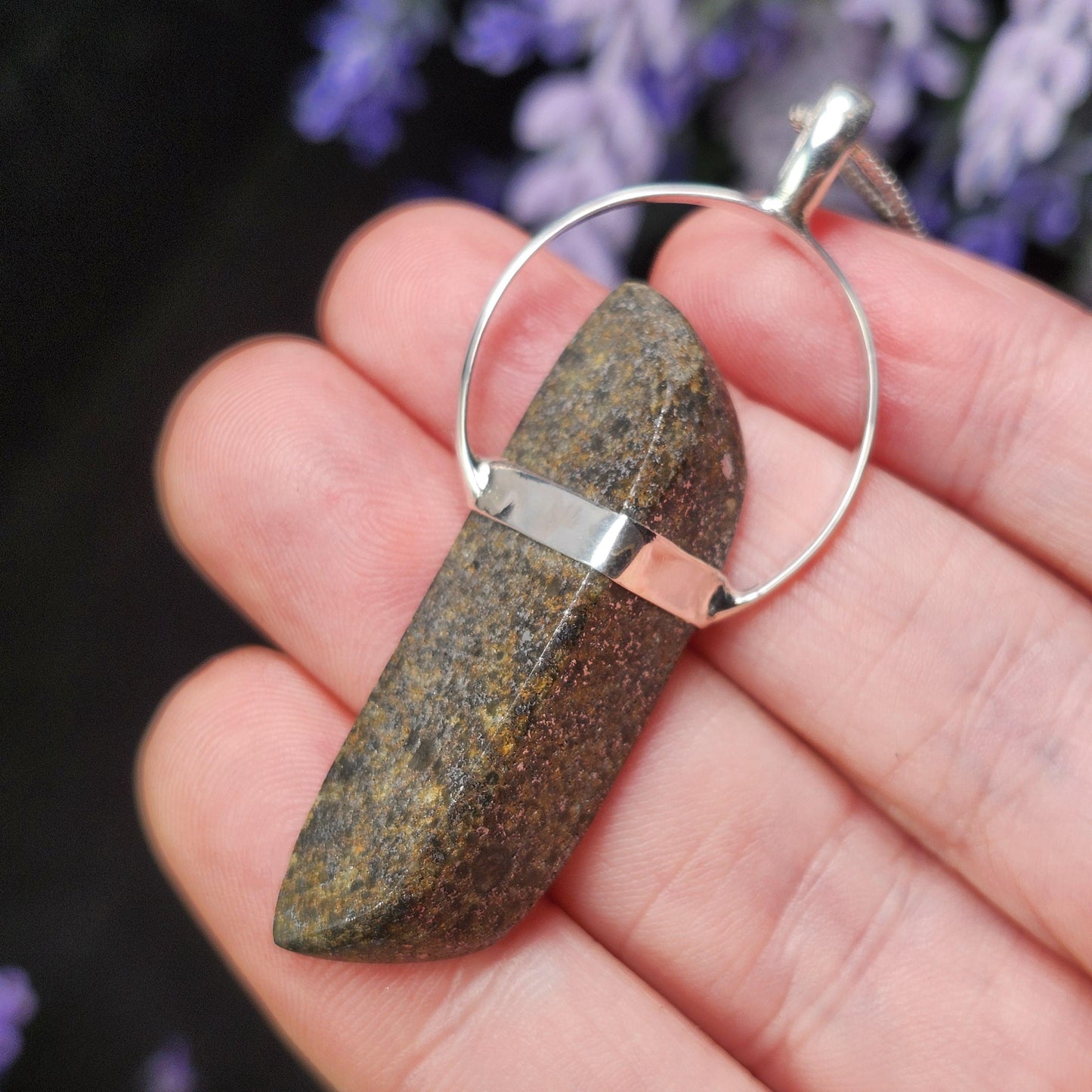 May include: A natural stone pendant with a silver-colored bail and a matching band. The stone is a dark brown color with lighter brown and yellow flecks. The pendant hangs from a silver-colored chain.