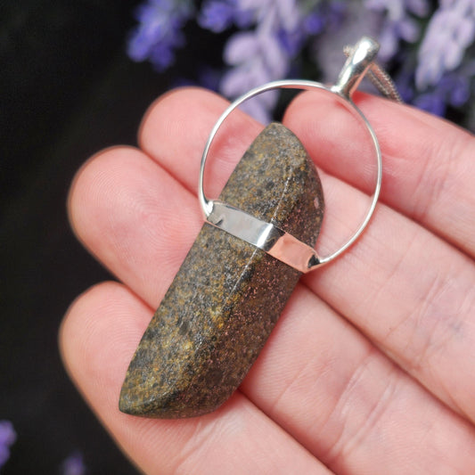May include: A natural stone pendant with a silver-colored bail and a matching band. The stone is a dark brown color with lighter brown and yellow flecks. The pendant hangs from a silver-colored chain.