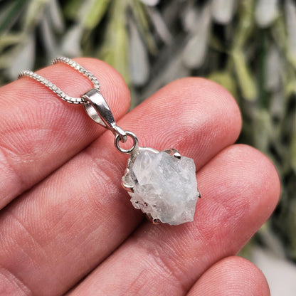 May include: A silver necklace featuring a raw, light blue crystal pendant. The pendant is irregularly shaped and held in a silver setting. The chain is a delicate, square-link style. The background is blurred, with green and white foliage.