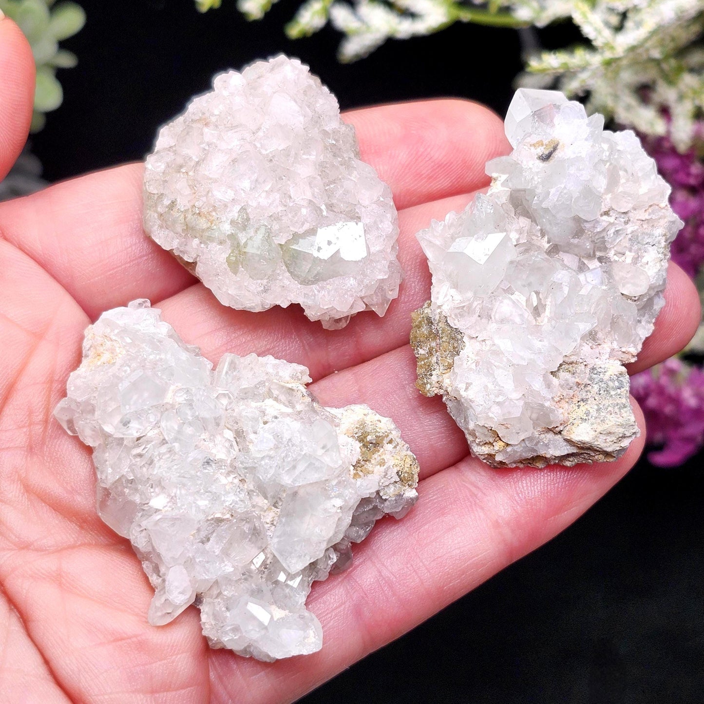 May include: Three clear quartz crystal clusters with a rough, textured appearance. The crystals are held in a hand, showcasing their natural, irregular shapes and sparkling facets. The quartz has a pale, translucent color.
